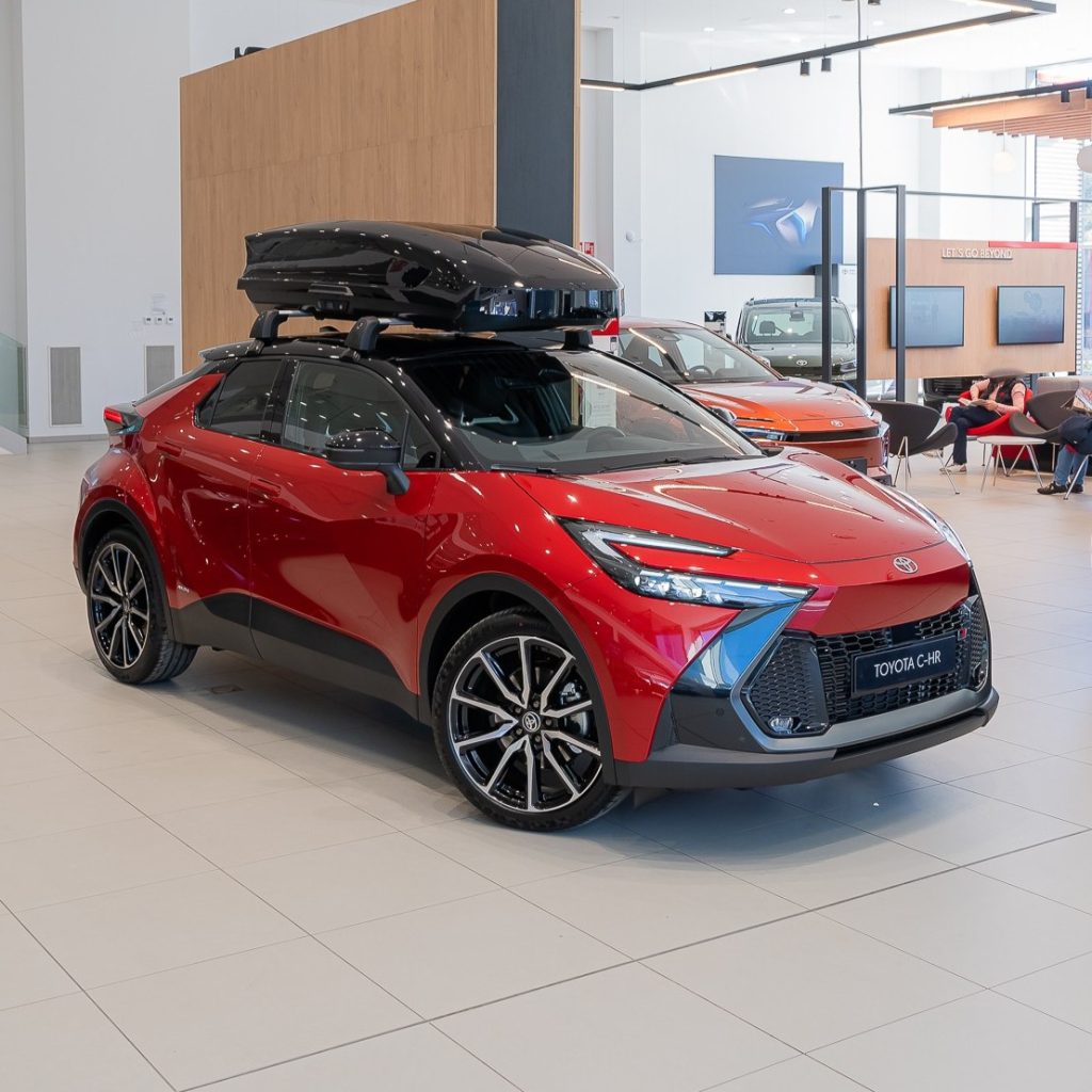 Red Toyota C-HR SUV on display in a bright showroom with a black roof cargo box mounted on top.