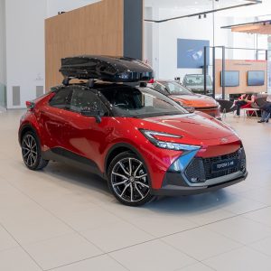 Red Toyota C-HR SUV on display in a bright showroom with a black roof cargo box mounted on top.