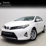 Front three-quarter view of a white Toyota sedan in a showroom, with 'Toyota Ocasión' banner in the background, highlighting the car's grille and headlights.