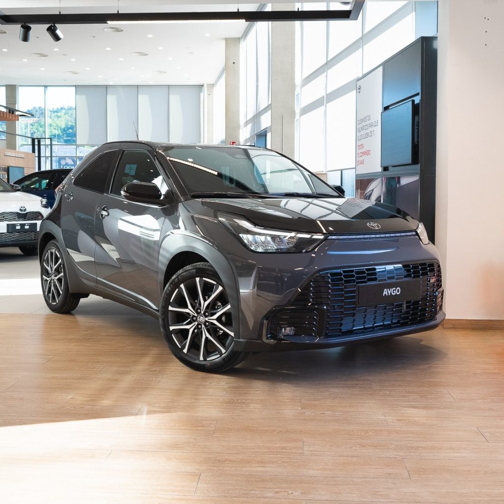 Grey Toyota Aygo compact hatchback on a car dealership showroom floor with large windows and other cars in the background